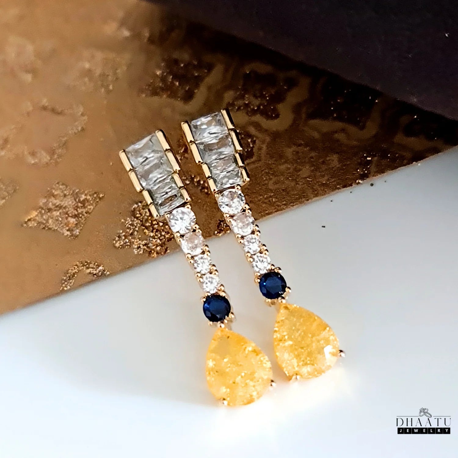 Gold-toned Zircon drop earrings with yellow and blue stones on a light-colored, textured surface